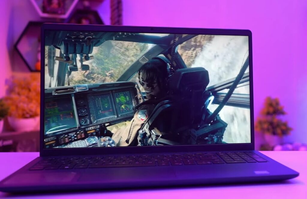 A Dell laptop around 40000 INR is shown on a desk with a vivid scene from a movie or video game displayed on its screen. The screen content features a person in a cockpit, wearing sunglasses and a headset, with various control panels and scenery visible in the background. The setting around the Dell laptop around 40000 has a soft, purple lighting, and there are some blurred decorative items in the background. The laptop's keyboard and touchpad are also visible, and the overall atmosphere appears to be designed for entertainment or gaming.