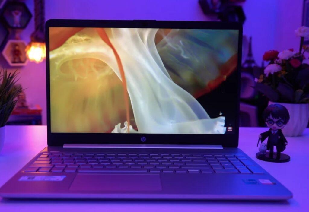 An HP laptop with a vibrant abstract image on the screen, set on a desk with purple and blue ambient lighting. The desk also features a small potted plant on the left and a Harry Potter figurine on the right.