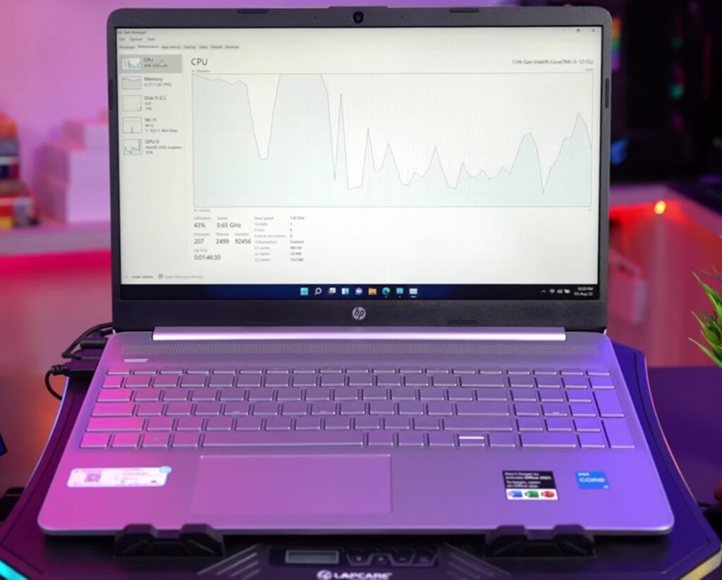 An HP laptop displaying the Task Manager on its screen, showing CPU usage and other system performance metrics. The laptop is placed on a cooling pad with a purple and blue ambient lighting setup in the background.