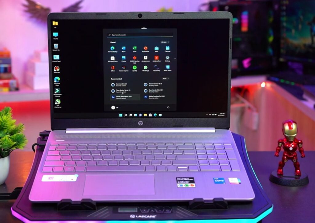 Front view of an HP laptop on a cooling pad, displaying the Windows Start menu. The laptop is set up on a desk with a small potted plant to the left and an Iron Man figurine to the right. The background features a blurred, colorful indoor setting with shelves and decorative items.