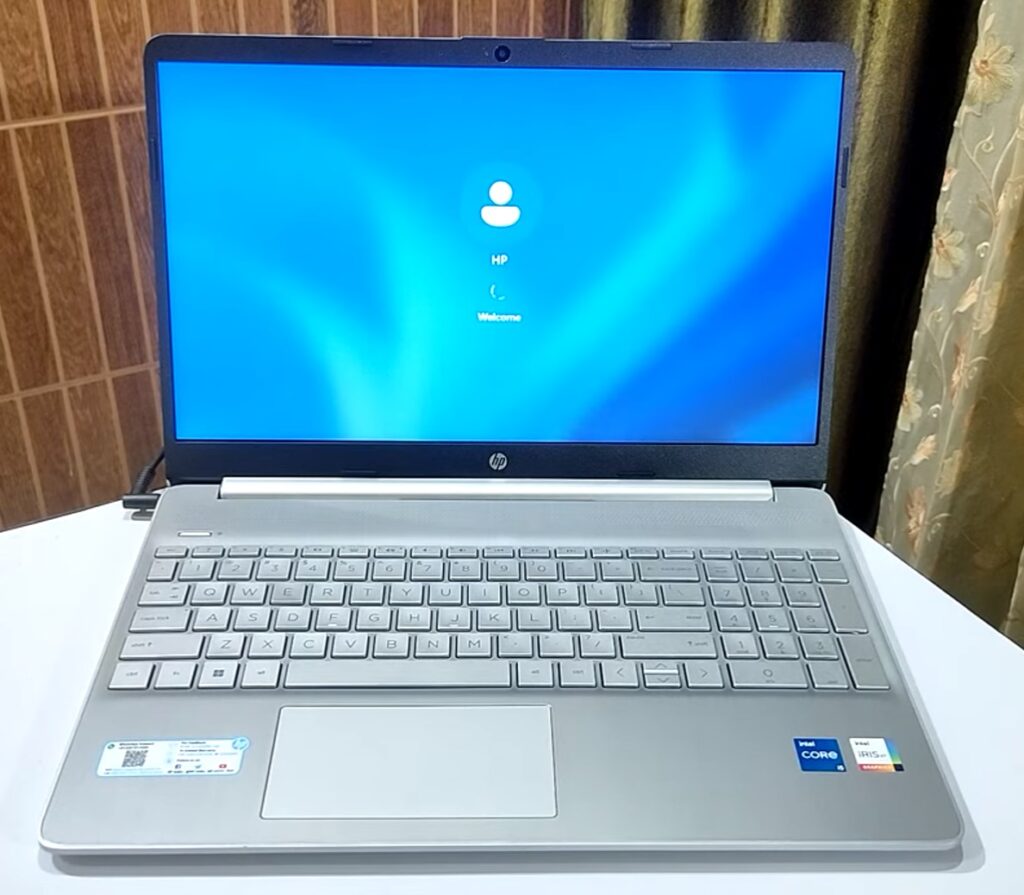 Front view of an HP laptop on a white table. The laptop features a silver chassis, a full-sized keyboard, and a trackpad. The screen displays a welcome login screen with the HP logo and user icon. Intel Core and Iris Xe graphics stickers are visible on the bottom right corner of the laptop.